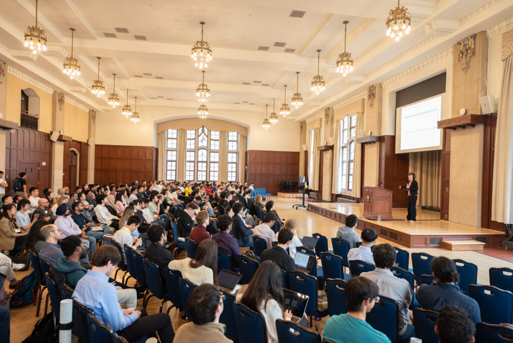 A woman stands on a stage in a ballroom, in front of a large seated audience.